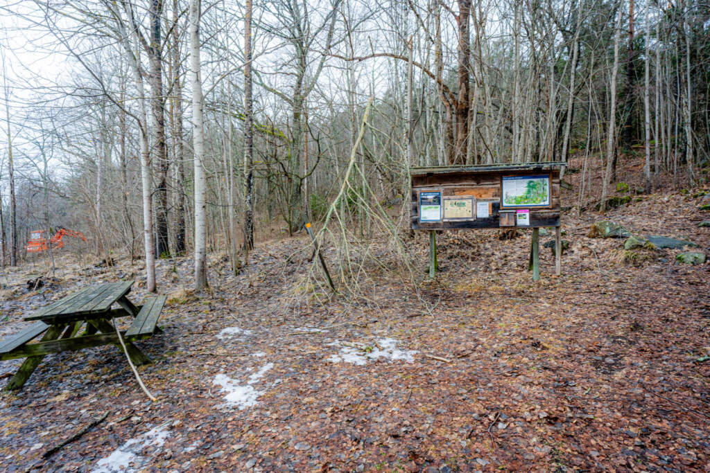 Informationstavla och sittbänk vid entrén till Kvillingeforkastningens naturreservat, lövskog utan löv i tidig vår med lite snö på marken.
