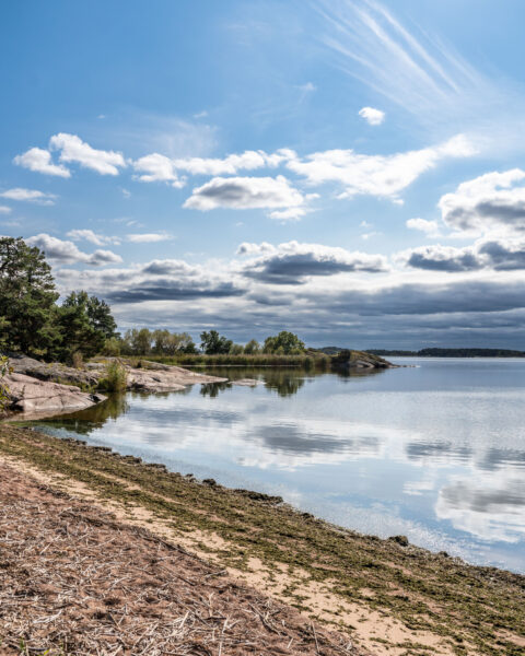 Stilla vik vid Grensholmsbadet med sandstrand, slipade klippor, grönska och spegelblank vattenyta under en molnströsslad sommarhimmel.