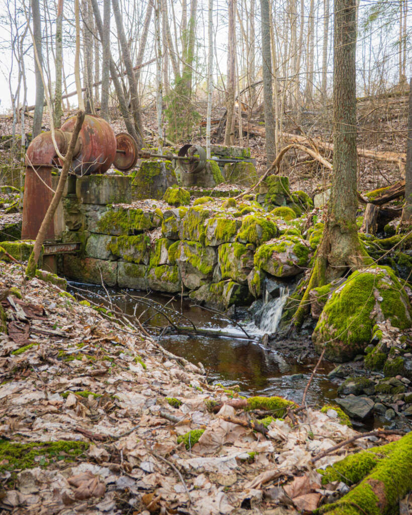 Mossbevuxna stenruiner från gammal kvarn vid Kvarseboån med rostig maskinrulle och porlande bäck i lövskog längs Kvarsebo kultur- och naturstig.