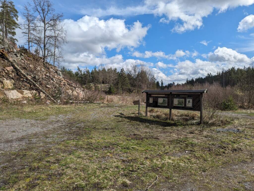 Informationstavla vid parkeringen till Hinnerstorp naturreservat med bergvägg, grusplan och barrskog under molnig vårhimmel.