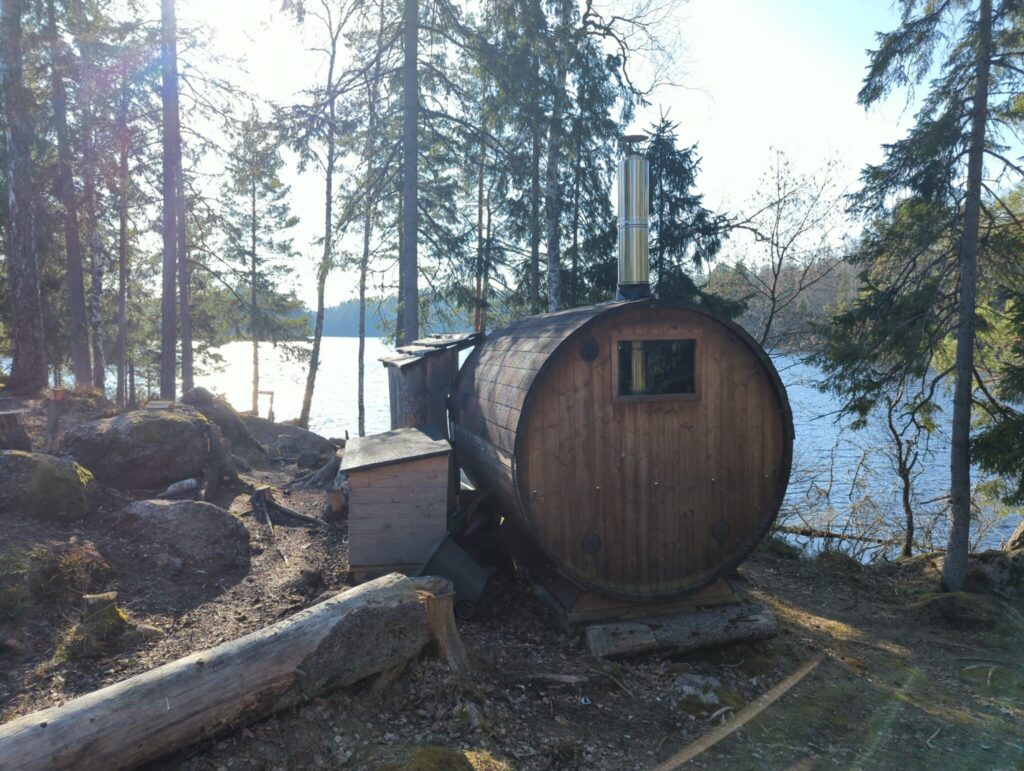 Vedeldad tunnbastu på en skogsklädd udde vid Övre Glottern i Norrköping, omgiven av barrträd och med glittrande sjövatten i bakgrunden.