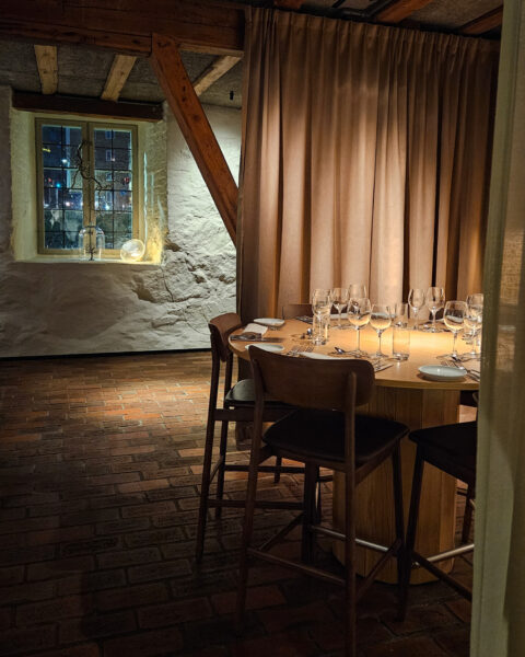 Stämningsfullt dukat bord med vinglas i en rustik restaurang med vitputsade stenväggar, träbjälkar och tegelgolv.