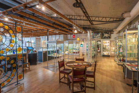 Utställningshall i Reijmyre Glasmuseum med glasmontrar, färgglatt blyglaskonstverk, hängande lampor och sittgrupp i trä.