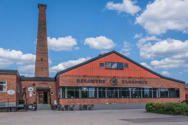 Reijmyre Glasbruk med tegelbyggnad, röd fasad och hög skorsten mot blå himmel i Finspångs kommun, Östergötland