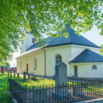 Regna kyrka i sommargrönska, vitputsad med mörkt tak, omgiven av kyrkogård med smidesjärnstaket och lummiga lövträd.