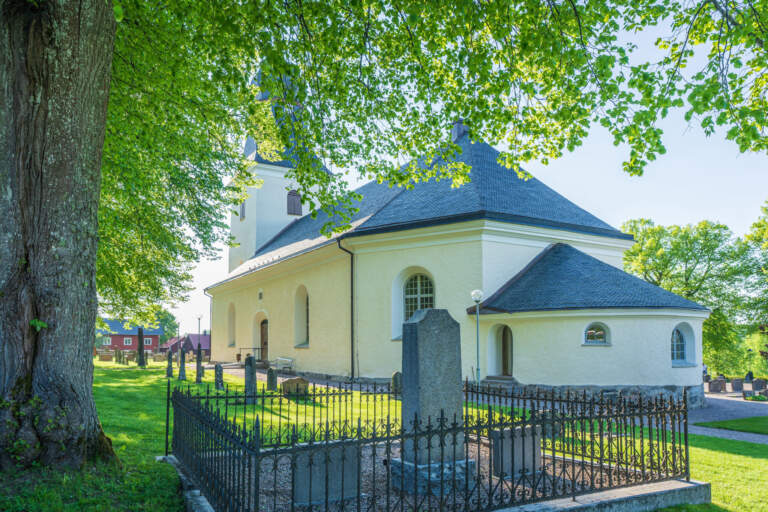 Regna kyrka i sommargrönska, vitputsad med mörkt tak, omgiven av kyrkogård med smidesjärnstaket och lummiga lövträd.