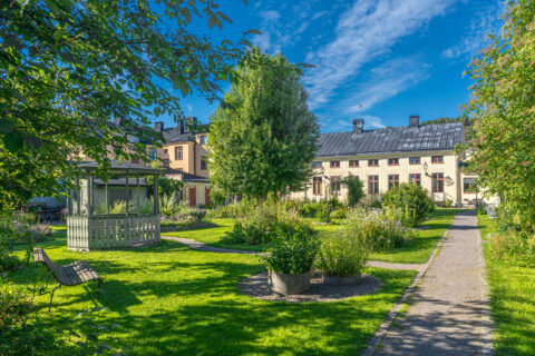 Grönskande Örtagården i Söderköping med grusgång, parkbänkar, blommande rabatter och historiska byggnader en solig sommardag
