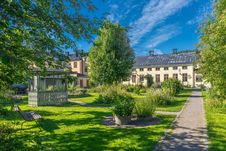 Grönskande Örtagården i Söderköping med grusgång, parkbänkar, blommande rabatter och historiska byggnader en solig sommardag