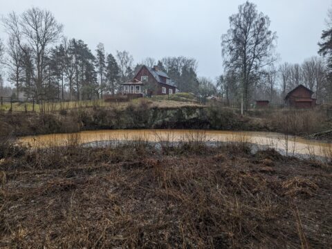 Vatenfylld gruvhåla vid Storgruvan i Hällestad, omgiven av vintrig vegetation med en röd stuga och uthus på höjden bakom.