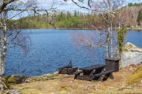 Grillplats med mörkt trä-bänkbord och eldstad vid en blå sjö i Öna kulturreservat, Linköpings kommun.
