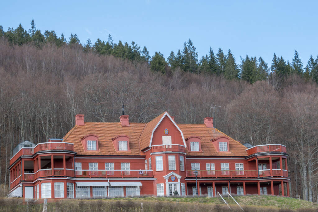 Ombergs Turisthotell, ett stort rött trähus med orange tegeltак och verandor, omgivet av lövskog och barrskog mot blå himmel.