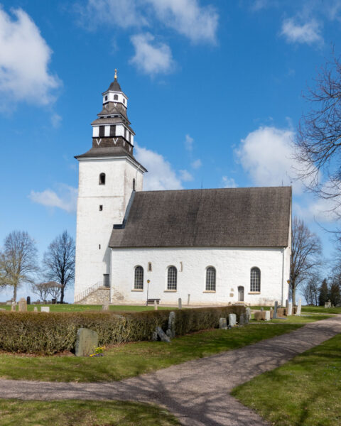 Vitkalkad Hovs kyrka med mörkt tak och karakteristiskt torn, omgiven av kyrkogård med häckar och gravstenar en vårdag i Vadstena kommun.