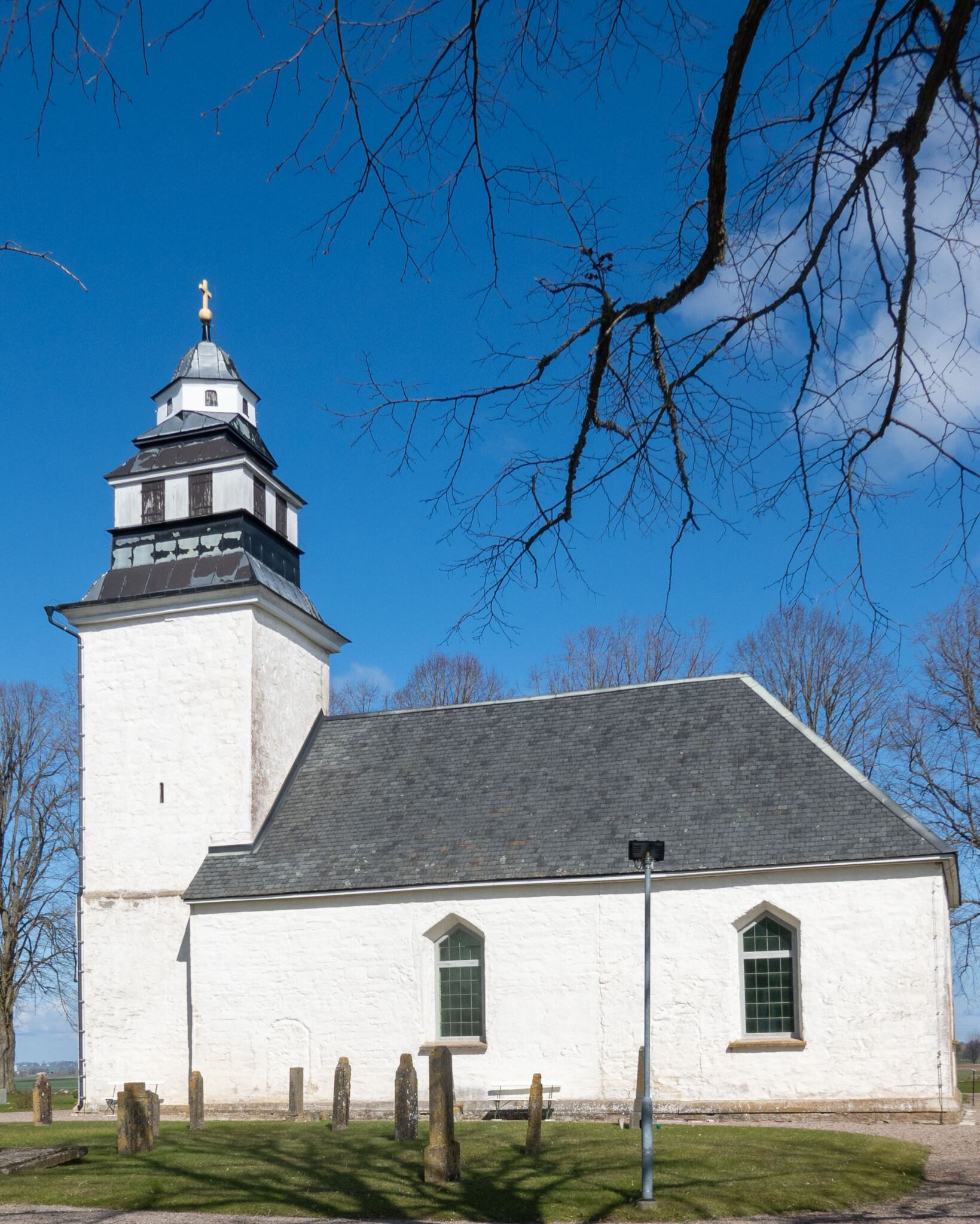 Vitputsade Strå kyrka med medeltida torn och skiffertak, omgiven av gravstenar och kala träd en klarblå vårdag nära Tåkern i Östergötland
