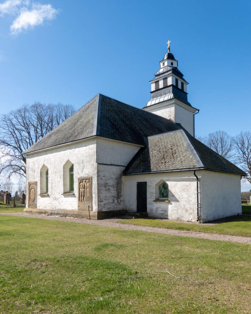 Vitputsad medeltida Strå kyrka med mörkt tak och klocktorn krönt av guldkors, omgiven av grön gräsmatta och kyrkogård nära Tåkern.