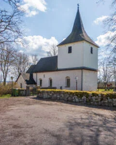 Väversunda kyrka med vitputsat långhus, stenmur och trätorn med spira, omgiven av kala träd en vårdag i Östergötland