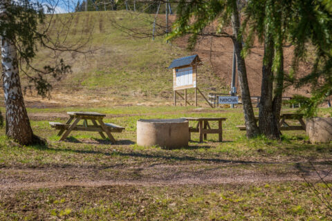 Rastplats med picknickbänkar och grillplats vid Ombergsliden naturreservat, med informationstavla och grön sluttning i bakgrunden.