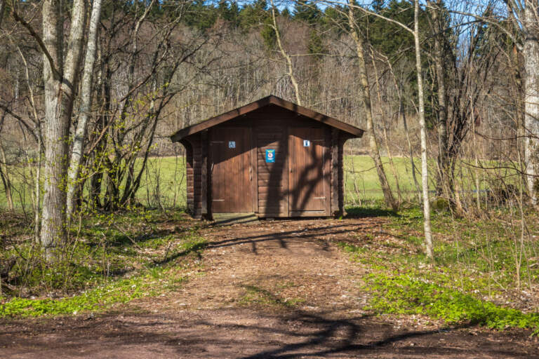 Brunmålat torrdass vid Höje Lilläng på Omberg, omgivet av lövträd och grönska en solig vårdag