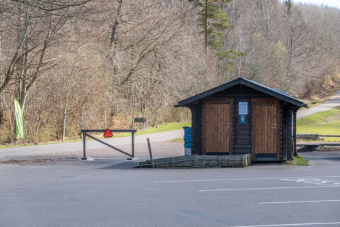 Toalettbyggnad i trä vid parkeringen på Omberg, Östergötland, omgiven av lövskog i tidig vår.