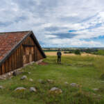 Person blickar ut över Östgötaslätten vid Sverkersgården på Omberg, intill en gammal träbyggnad med tegeltak och gröna ängar.