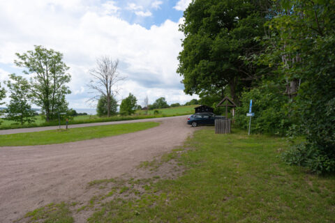 Grusad parkering vid Almelyckan på Ombergs södra sida med en parkerad bil, informationsskylt, gröna ängar och lummiga träd en sommardag.