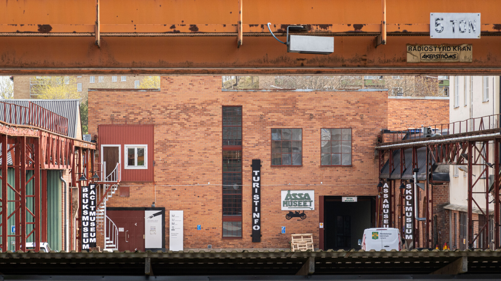 Tegelbyggnad med skyltar för ÅSSA Museet, Turistinfo, Bruksmuseum, Facitmuseum och Skolmuseum i Åtvidaberg, sedd under en industrikran