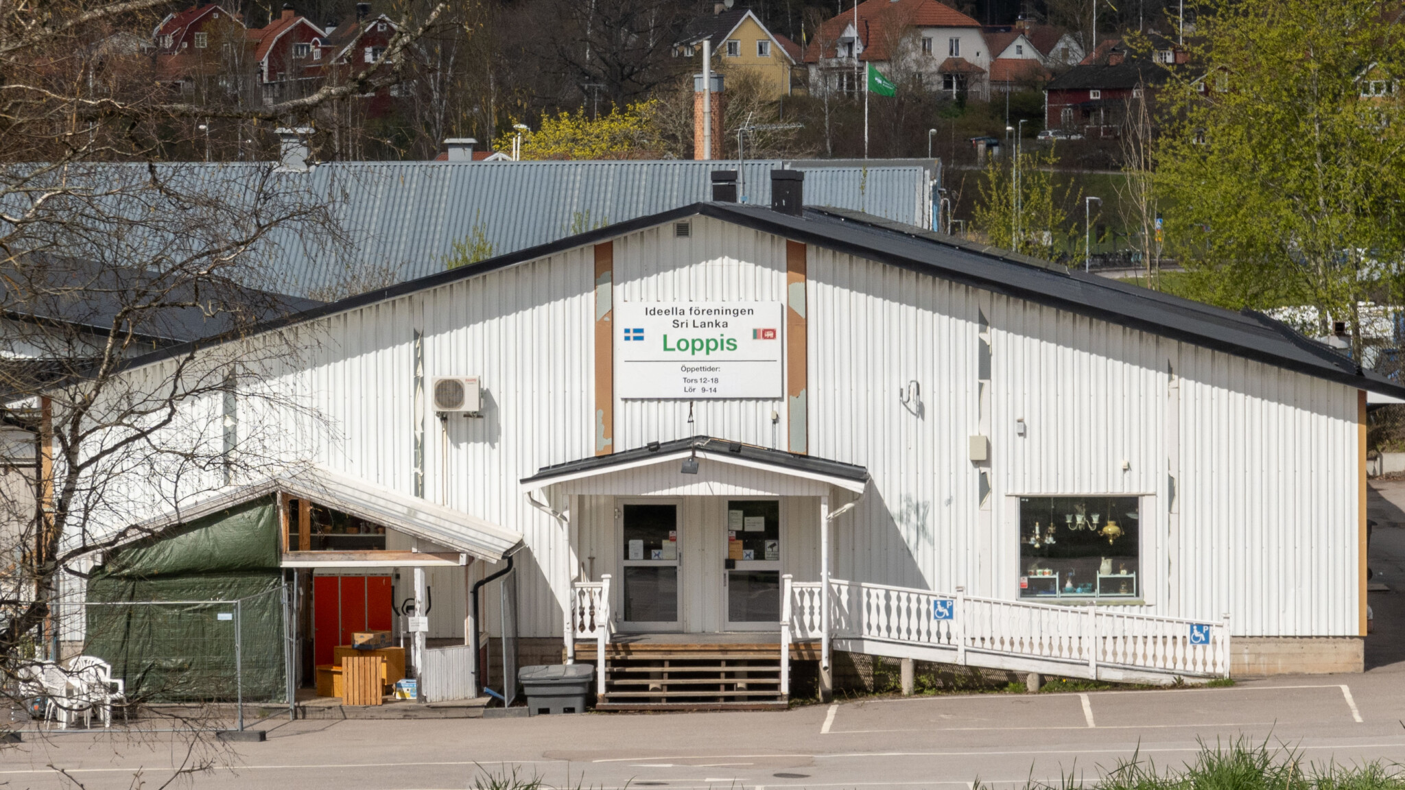 Vit plåtbyggnad med skylt "Loppis" tillhörande ideella föreningen Sri Lanka i Åtvidaberg, med veranda och bostadshus i bakgrunden.