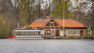 Röd träbyggnad med oranget tegeltak vid Bysjön i Åtvidaberg, med musikbåten och signalflaggor vid bryggan en vårdag