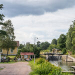 Brådtom sluss längs Göta kanal med vattenfall över slussporten, gult hus med sommarcafé och besökare vid bänkar, omgivet av grönska.