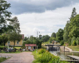 Brådtom sluss längs Göta kanal med vattenfall över slussporten, gult hus med sommarcafé och besökare vid bänkar, omgivet av grönska.