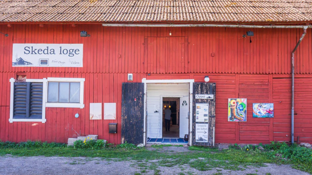 Röd ladugård med skylten "Skeda loge", öppna portar som välkomnar besökare till konstutställningar och hantverk i Östergötland