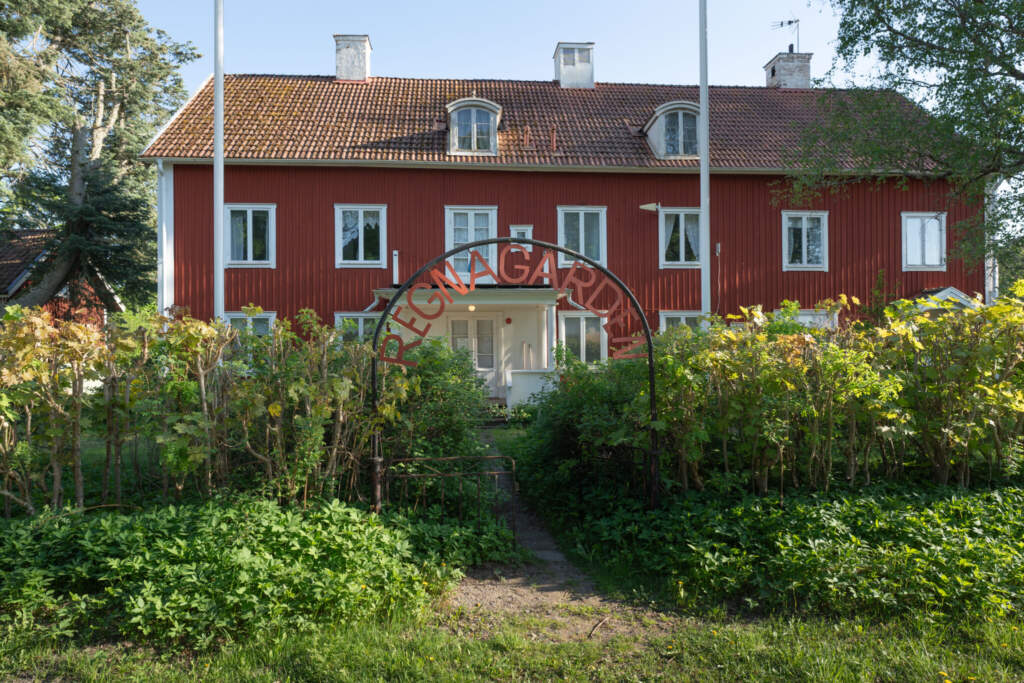 Röd tvåvånings träbyggnad med vita knutar vid Regnagården, omgiven av frodig trädgård med portal i smidesjärn, Regna Vandrarhem i Östergötland.