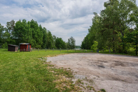 Grusad parkering vid Bankekinds badplats med gröna gräsytor, liten röd stuga och lummiga lövträd en sommardag med molnig himmel.
