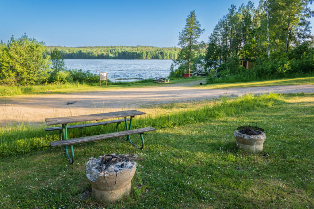 Grillplatser och sittbänk vid Byrumsbadet med utsikt över sjön, brygga och grönskande skog en solig sommardag i Åtvidaberg