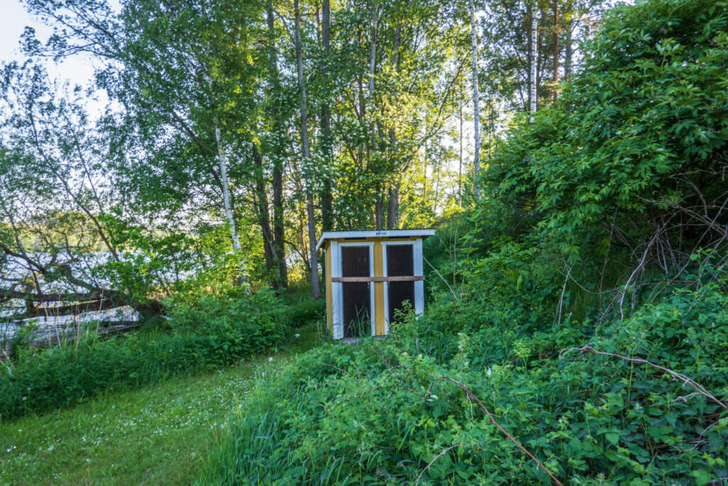 Litet omklädningshytt i grönska vid Byrumsbadet, omgiven av lövträd och frodig vegetation nära vattnet i Åtvidaberg.