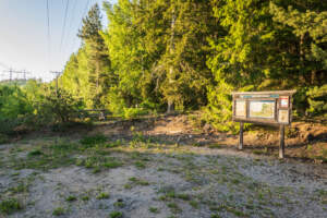Parkering vid Ristskeda naturreservat med informationstavla, sittbänk och grusplan omgiven av barrskog i sommargrönska.