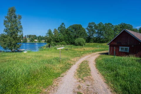 Grusväg genom grönt landskap vid Sätravallen med röd stuga, sjö med badbrygga och lummiga träd en solig sommardag