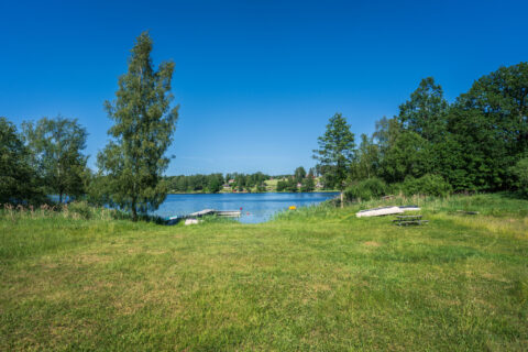 Grönklädd strandäng vid Sätravallens badplats med badbrygga, uppdragna båtar och picknickbänk vid en stilla sjö en solig sommardag