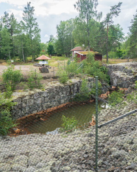 Vattenfyllt gruvhål vid Mormorsgruvan i Åtvidaberg, omgärdat av stängsel och bergväggar, med rastplats och röd stuga i bakgrunden