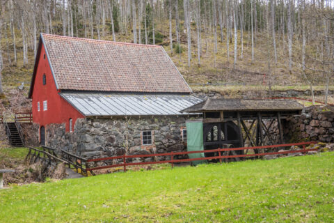 Röd kvarnbyggnad med stenmur och tegeltak vid Forsaström, omgiven av grön äng och lövskog längs Östgötaleden.