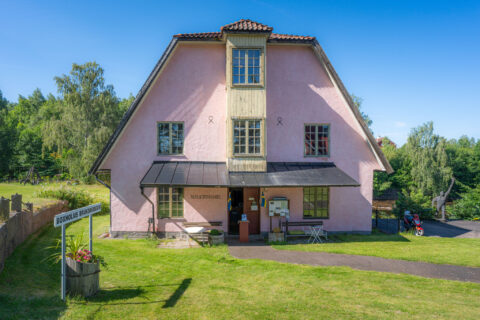 Rosa kvarnbyggnad med mansardtak som inrymmer Boxholms Bruksmuseum, omgiven av grön gräsmatta och lummiga träd en solig sommardag.