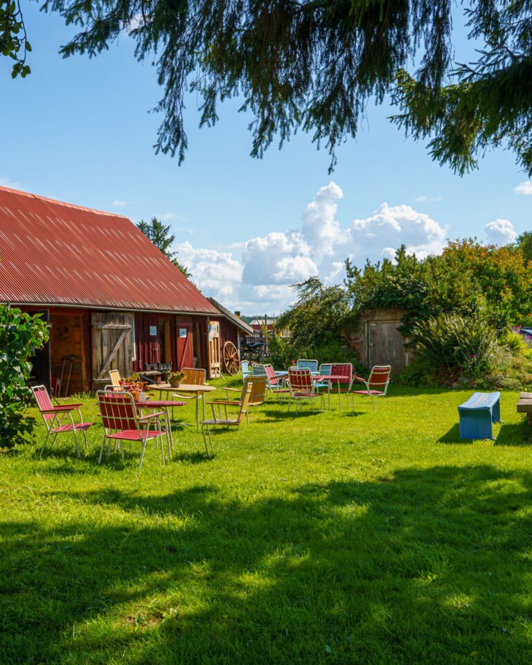 Idyllisk trädgård med retrostolar på grön gräsmatta vid röd lada med plåttak hos Botanicus Gårdsbutik i Mjölby, en solig sommardag