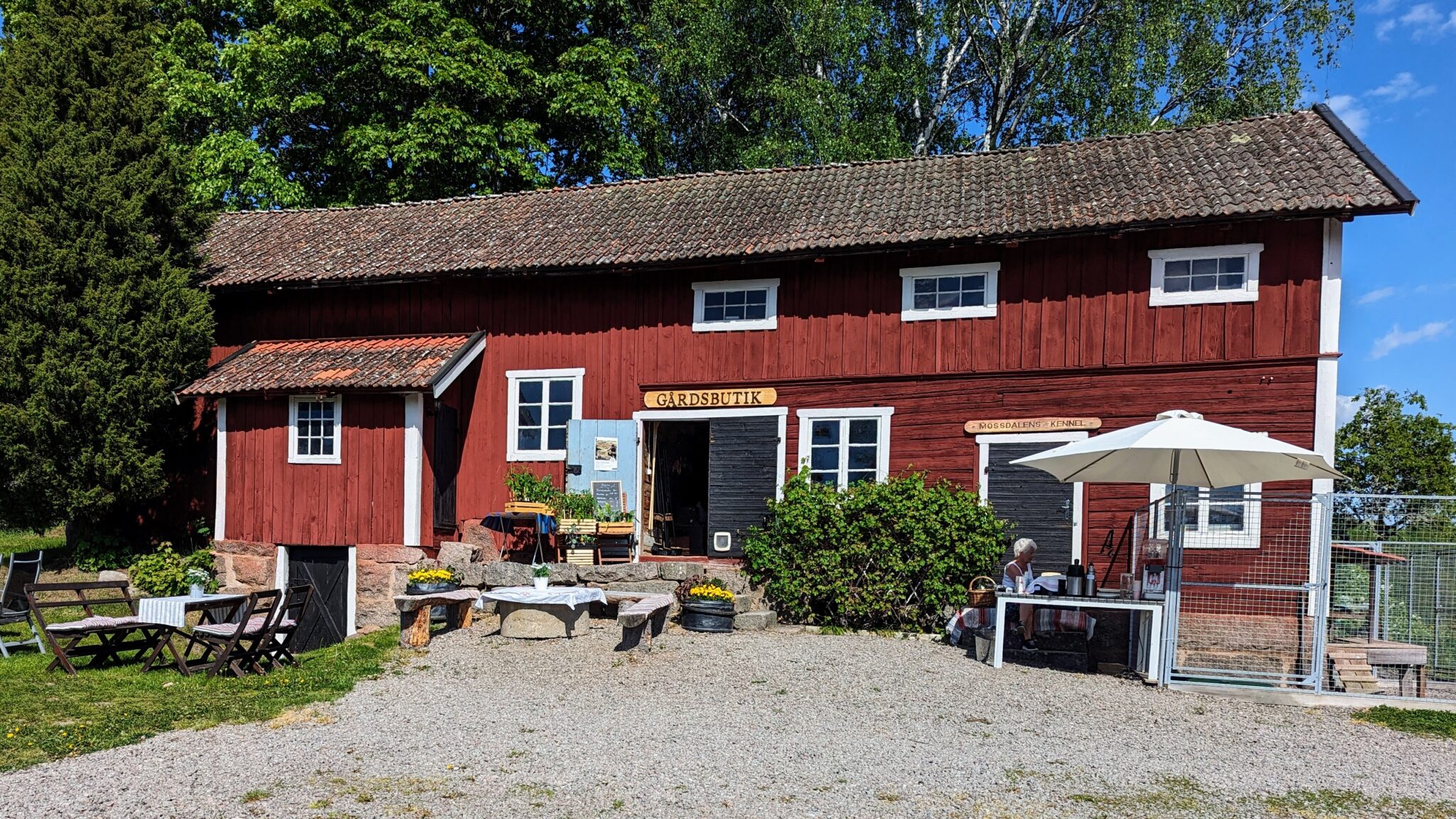 Röd timmerbyggnad med skylten "Gårdsbutik" i soligt sommarväder, omgiven av grönskande träd i Kinda, Östergötland.