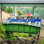 Passagerarbåten Dalslandia passerar akvedukten i Håverud med flaggspel, medan besökare promenerar på den gröna akvedukten nedanför järnvägsbron.