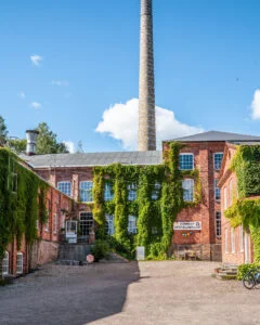 Murgrönsklädda tegelbyggnader med hög fabriksskorsten vid Not Quite i Fengersfors, med skylt för sommarutställningar en solig dag