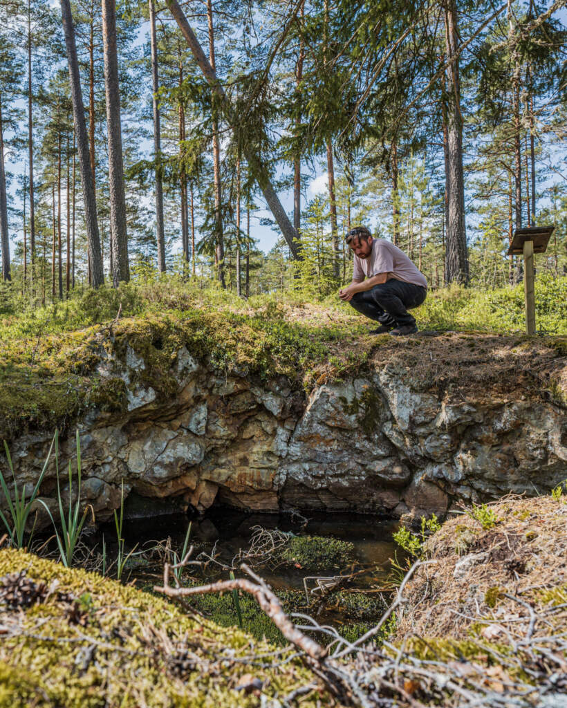 Man sitter på huk vid kanten av ett gammalt gruvschakt fyllt med vatten, omgivet av mossbeklädd berggrund och tallskog i Hällestad, Östergötland