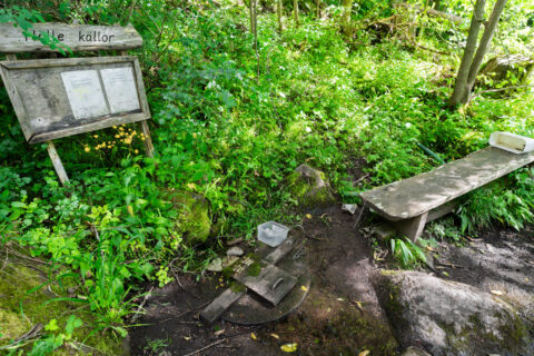 Hälle källor på Omberg – en naturkälla omgiven av frodig grönska, med informationsskylt, träbänk och vattenuppsamling vid källan.
