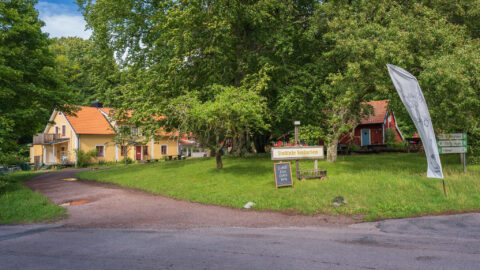 Stocklycke Vandrarhem på Omberg med gult hus, röd stuga och välkomstskylt omgivna av lummiga lövträd en sommardag