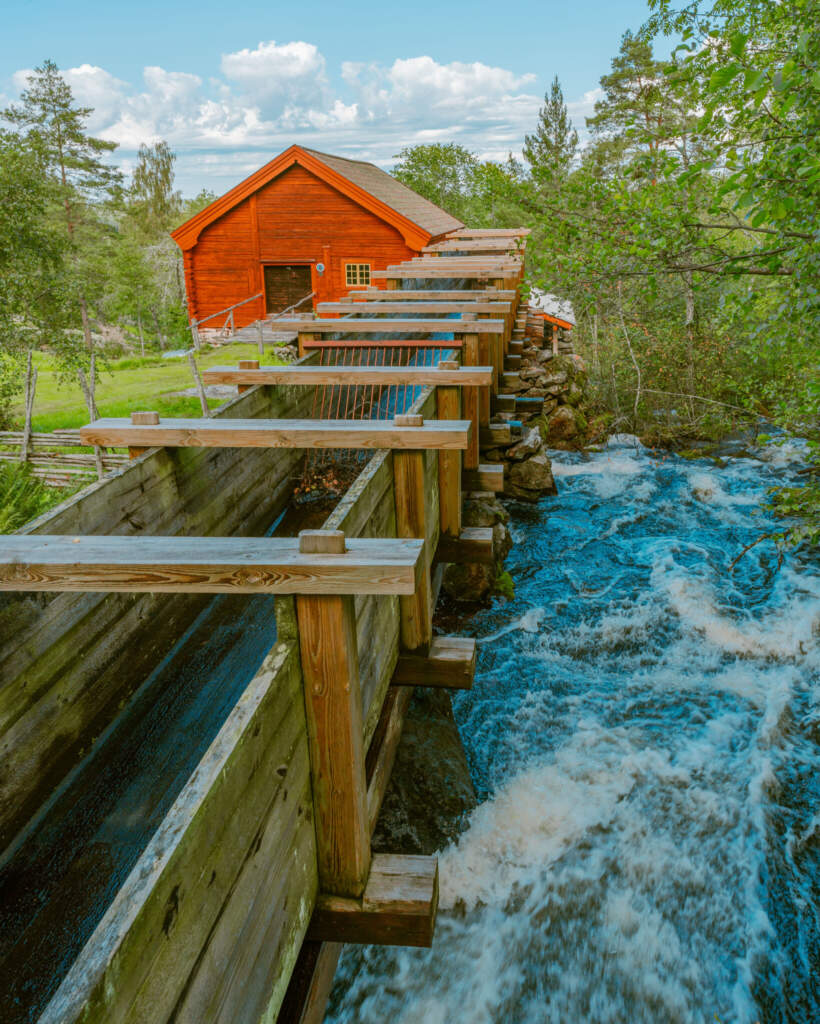 Röd 1700-talskvarn vid Råås kvarn i Ydre med träränna och forsande vatten, omgiven av grönska en sommardag