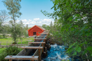 Rödfärgade Råås kvarn i Ydre med träränna för vatten längs en forsande bäck, omgiven av grönska och gärdesgård en sommardag