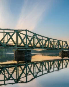 Grön stålbro över Dalslands kanal vid solnedgång, med spegelblank vattenyta som reflekterar brokonstruktionen, nära Bengtsfors